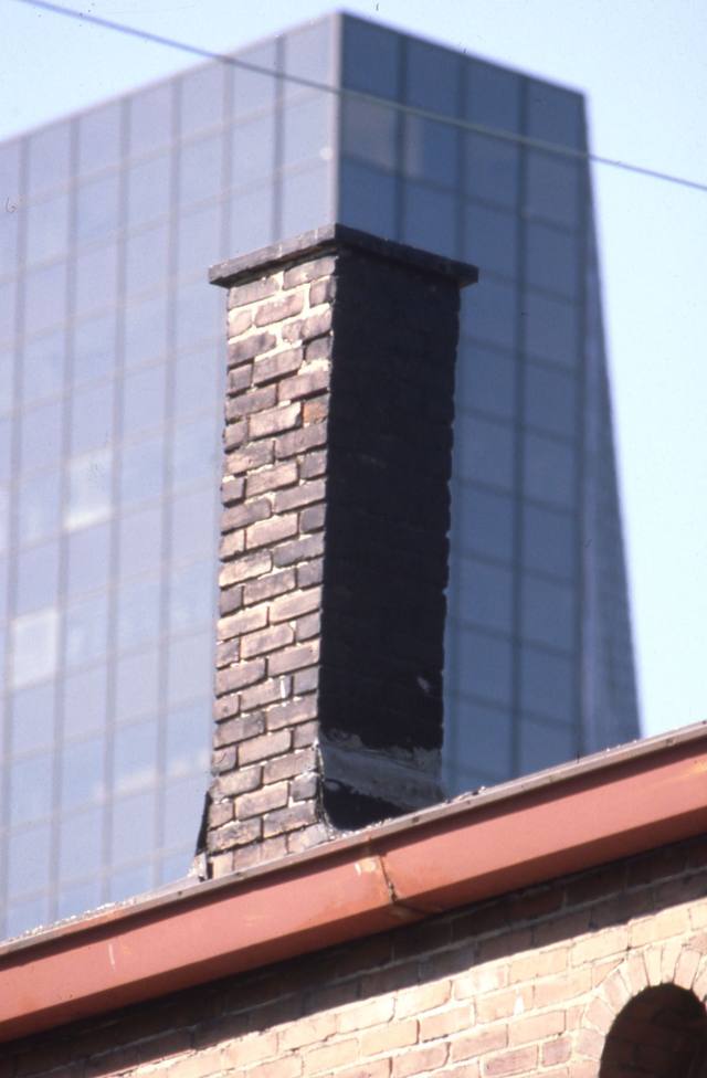 Weathered brick chimney with a concrete cap in front of a glass tower